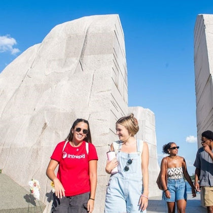 People walking near large stone structures on a sunny day, one wearing a red shirt and another in light denim overalls.