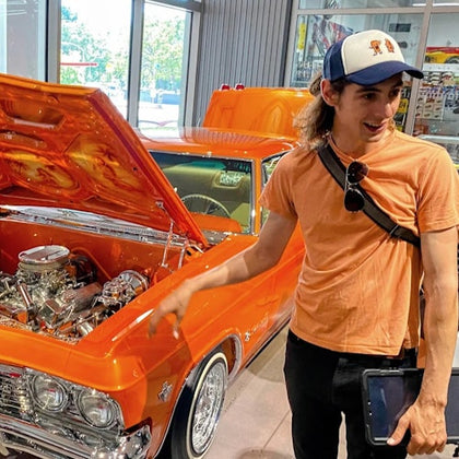 A person in an orange shirt and baseball cap stands excitedly near an orange classic car with its hood open indoors.