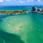 Aerial view of numerous boats anchored in turquoise water near a bridge and high-rise buildings with the ocean in the background.