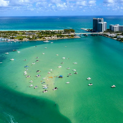 Aerial view of numerous boats anchored in turquoise water near a bridge and high-rise buildings with the ocean in the background.
