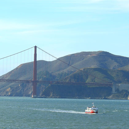 A boat sails on water with the Golden Gate Bridge and a mountain in the background under a clear blue sky.