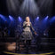 A singer performs on stage under dramatic lighting, surrounded by musicians in stylized costumes.