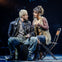 Two actors in costume perform on stage; one wearing rugged attire, the other in a layered bohemian outfit, seated on chairs.