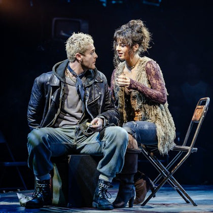 Two actors in costume perform on stage; one wearing rugged attire, the other in a layered bohemian outfit, seated on chairs.