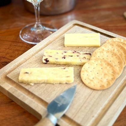 A wooden board with slices of cheese, crackers, and a knife on it, with a wine glass in the background.