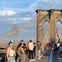 People walking and taking photos on the Brooklyn Bridge under a blue sky, with the American flag in the background.