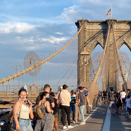 People walking and taking photos on the Brooklyn Bridge under a blue sky, with the American flag in the background.