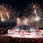 A nighttime stage performance with bright fireworks in the sky, a globe structure on stage, and a large audience watching.
