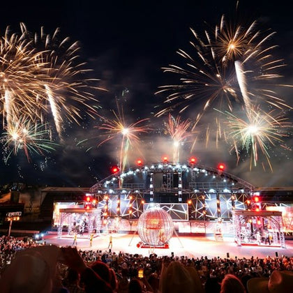 A nighttime stage performance with bright fireworks in the sky, a globe structure on stage, and a large audience watching.