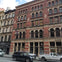 A red-brick building with arched windows and intricate details, housing shops on the ground floor, with cars parked outside.