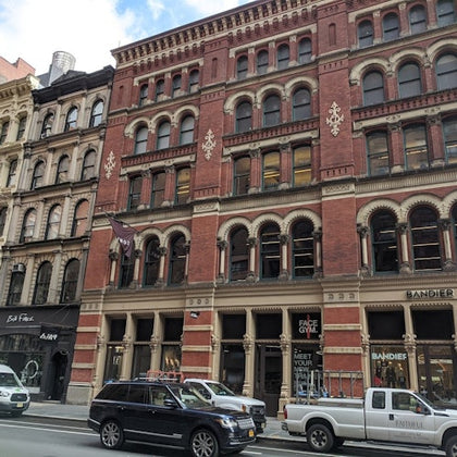 A red-brick building with arched windows and intricate details, housing shops on the ground floor, with cars parked outside.