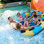 A group of people enjoying a water ride on a large inflatable raft in a pool with splashing water and a slide visible.