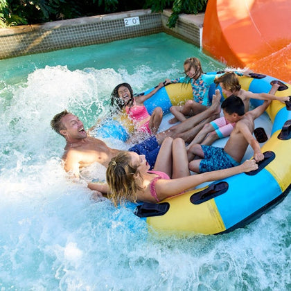 A group of people enjoying a water ride on a large inflatable raft in a pool with splashing water and a slide visible.