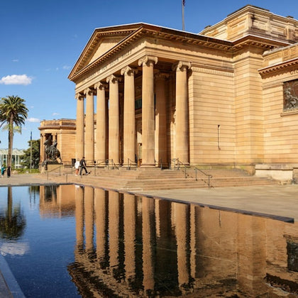 A neoclassical building with tall columns, reflected in a tranquil pool. Two people walk near the entrance. Palm trees and a blue sky are visible.