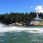 A white lighthouse on rocky shore surrounded by trees under a clear blue sky, with waves splashing in the foreground.