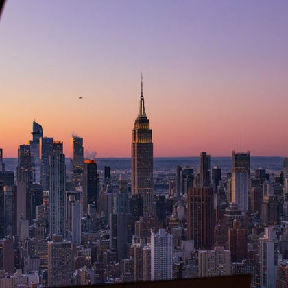 A cityscape at sunset featuring the Empire State Building and surrounding skyscrapers under a colorful sky.