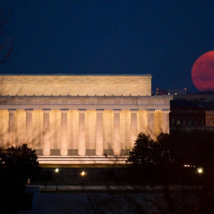 The Lincoln Memorial illuminated at night with a large red moon rising in the background.