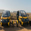 Two small, yellow and black electric cars parked on a dirt area near a coastline with hills in the background.