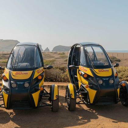 Two small, yellow and black electric cars parked on a dirt area near a coastline with hills in the background.