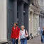 Two people walking on a sidewalk in front of a row of ornate buildings, one wearing a red jacket and the other a pink and blue jacket.