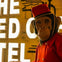 A person wearing a monkey mask and a red bellhop uniform stands in front of a lit sign reading 'The Ned Hotel.'