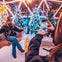 A person takes a photo of another person seated in a festive setting with decorated trees and bright lights.