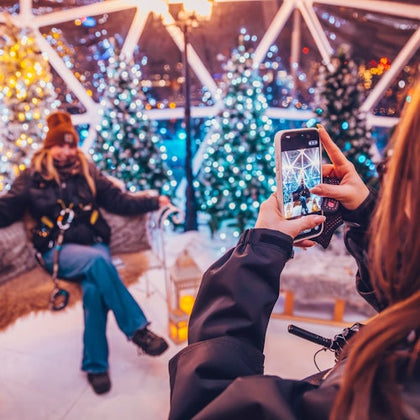 A person takes a photo of another person seated in a festive setting with decorated trees and bright lights.
