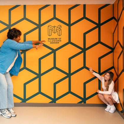 Two individuals interact with a perspective illusion wall at the Museum of Illusions in Denver, featuring a geometric pattern.