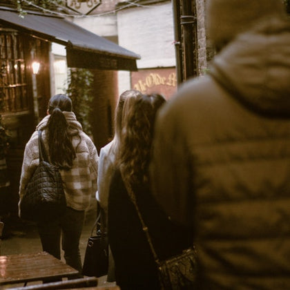 A group of people walking down an outdoor alleyway, with tables on the side, near a building with lit windows.