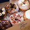 A box containing six assorted gourmet doughnuts, each topped with different glazes, crumbs, and toppings.
