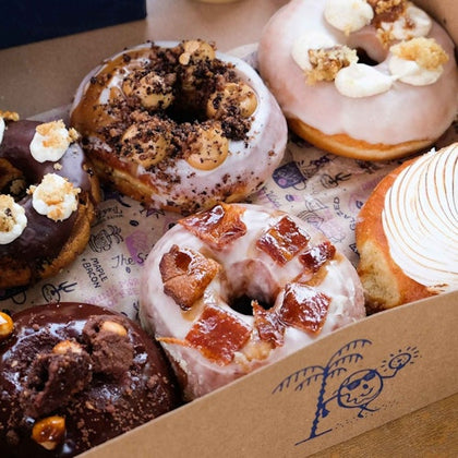 A box containing six assorted gourmet doughnuts, each topped with different glazes, crumbs, and toppings.