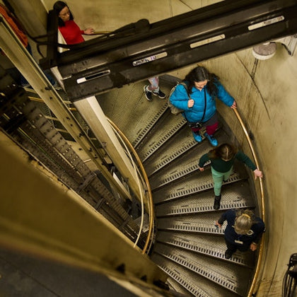 Several people descend a spiral staircase in an industrial setting with metal steps and a concrete wall.