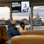 People seated on a ferry with a view of a warship and cityscape through the windows, and a screen displaying a message.
