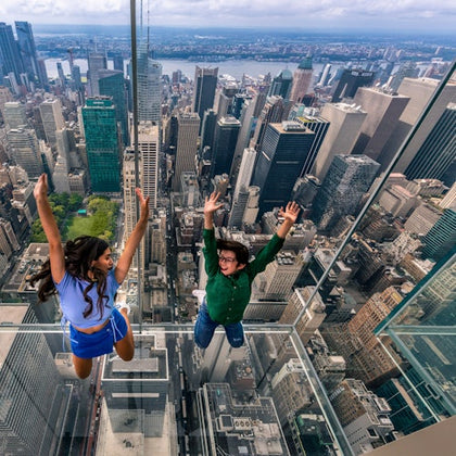 Two people on a glass floor at a high-altitude observation deck with a cityscape view in the background.