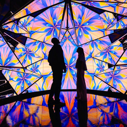 Two silhouetted figures standing inside a brightly lit, kaleidoscope-patterned room with reflective flooring.