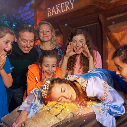 Six people gathered around a person dressed as Sleeping Beauty, who is lying on a rustic bed. A "Bakery" sign is visible behind them.