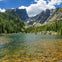 A serene mountain lake surrounded by evergreen trees and rocky peaks under a blue sky with scattered clouds.