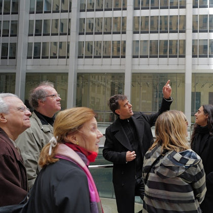 A group of people outside a modern building, with one man pointing upwards while speaking to the others.