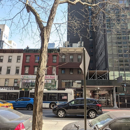 City street with parked cars, a bus, and buildings in various styles. A bare tree trunk and a sign are in the foreground.