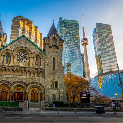 A historic stone church with arched doors and windows stands in front of modern skyscrapers, including a tower with a CN Tower.