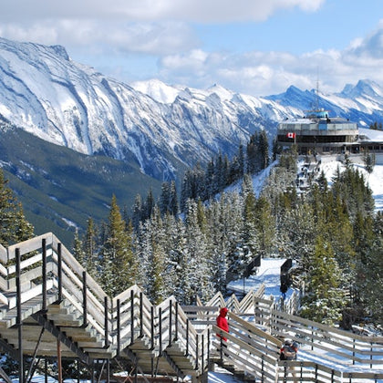 Snow-covered mountain view with a wooden stairway leading to a distant building with a Canadian flag. People wearing winter clothing.