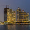 A waterfront cityscape at twilight showing lit high-rise buildings, a construction crane, and reflections on the water.