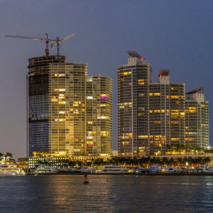 A waterfront cityscape at twilight showing lit high-rise buildings, a construction crane, and reflections on the water.