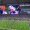 A football game at AT&T Stadium with a large screen displaying players in white and blue uniforms featuring star logos.