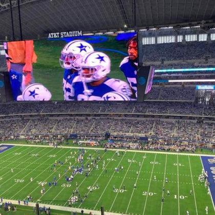 A football game at AT&T Stadium with a large screen displaying players in white and blue uniforms featuring star logos.