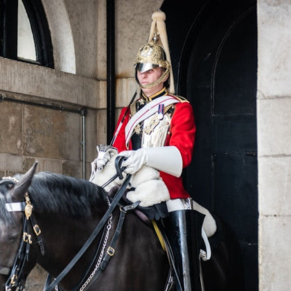 A guard in a red uniform and gold helmet sits on a black horse at an archway, holding reins with gloved hands.
