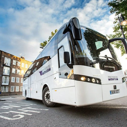 A white coach bus parked on a street with brick buildings and trees in the background, under a partly cloudy sky.