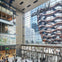 Shops and people in a modern, multi-story mall with large windows reflecting an intricate, honeycomb-like outdoor structure.