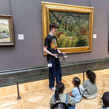 A person giving a presentation to a group of seated children in front of a large, framed painting in an art gallery.