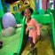 A child wearing a pink dress with a rainbow design plays on a green slide in a colorful indoor play area with large props.